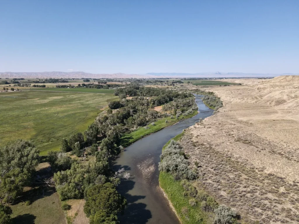 Aerial view of a river winding through a landscape featuring green agricultural fields on the left and arid, rocky terrain on the right, with distant mountains under a clear blue sky.