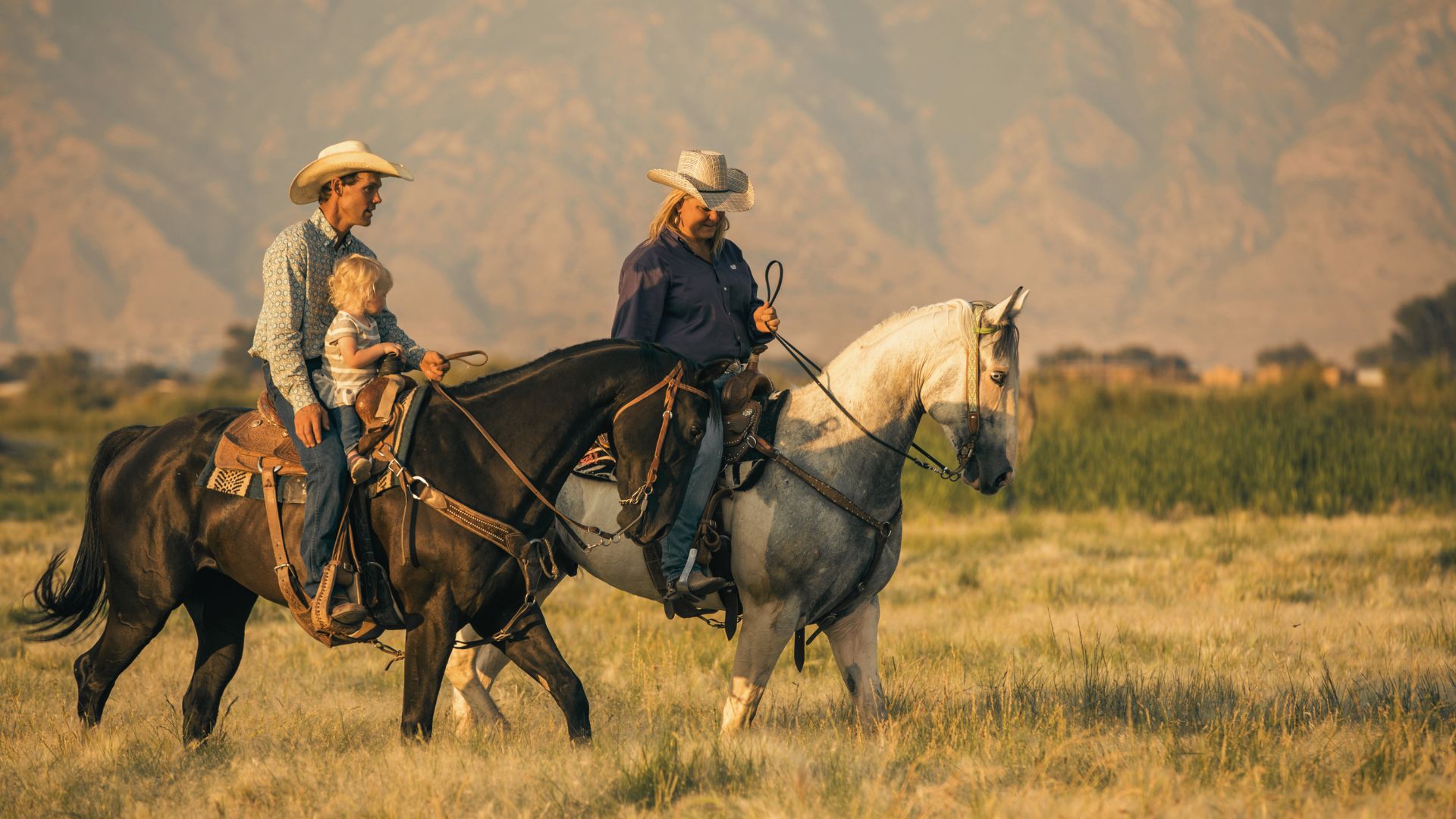A man, woman, and young child ride horses in a grassy field with mountains in the background. The man, wearing a white hat and plaid shirt, holds the child on a dark horse. The woman, in a blue shirt and white hat, rides a white horse beside them as they discuss their private credit options for financing the ranch.